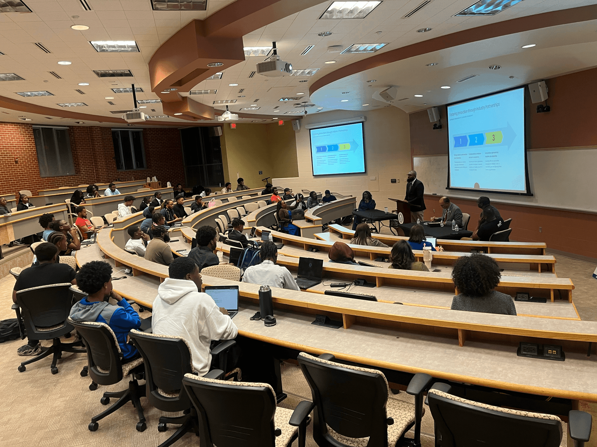 Classroom with students and a panel discussion taking place, presentation showing on screens.