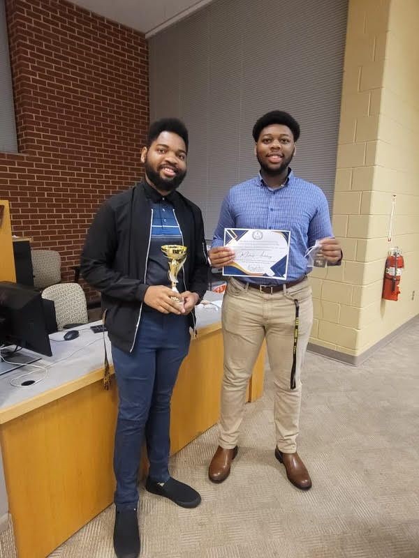 Two men standing in a room, one holding a trophy and the other holding a certificate.