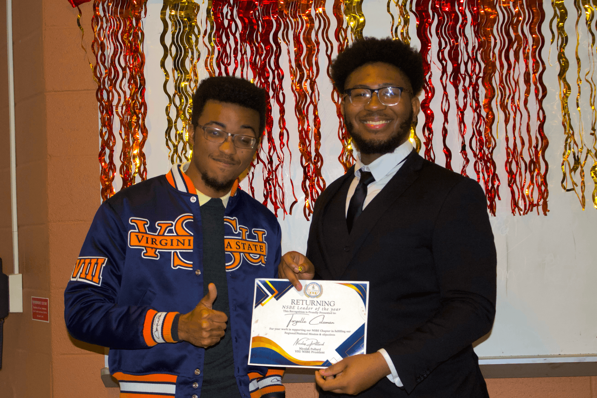 Two men smiling, one holding a certificate, in front of red and gold streamers.