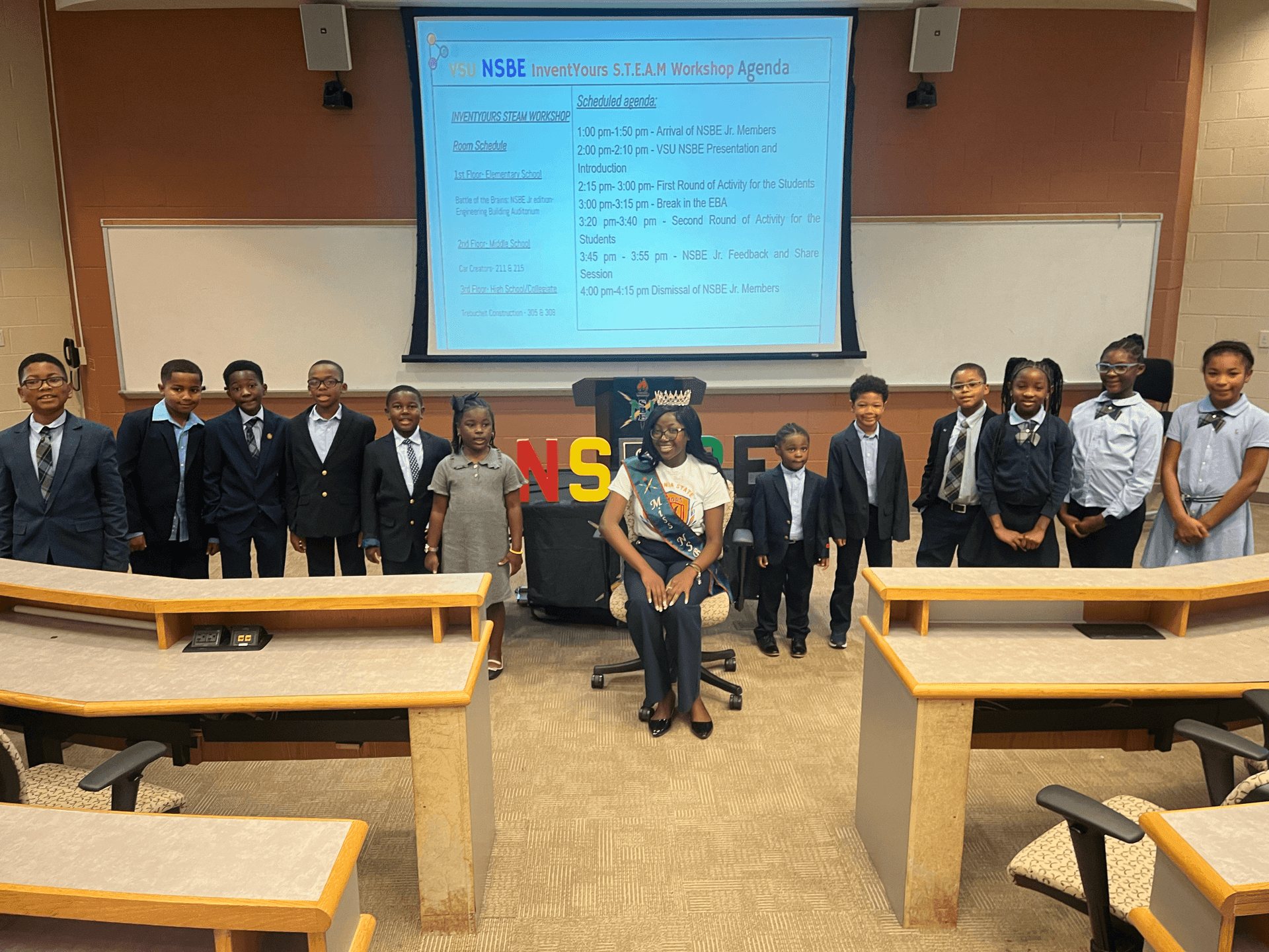 Group of children in formal attire standing in a classroom with a presentation screen displaying an event agenda.