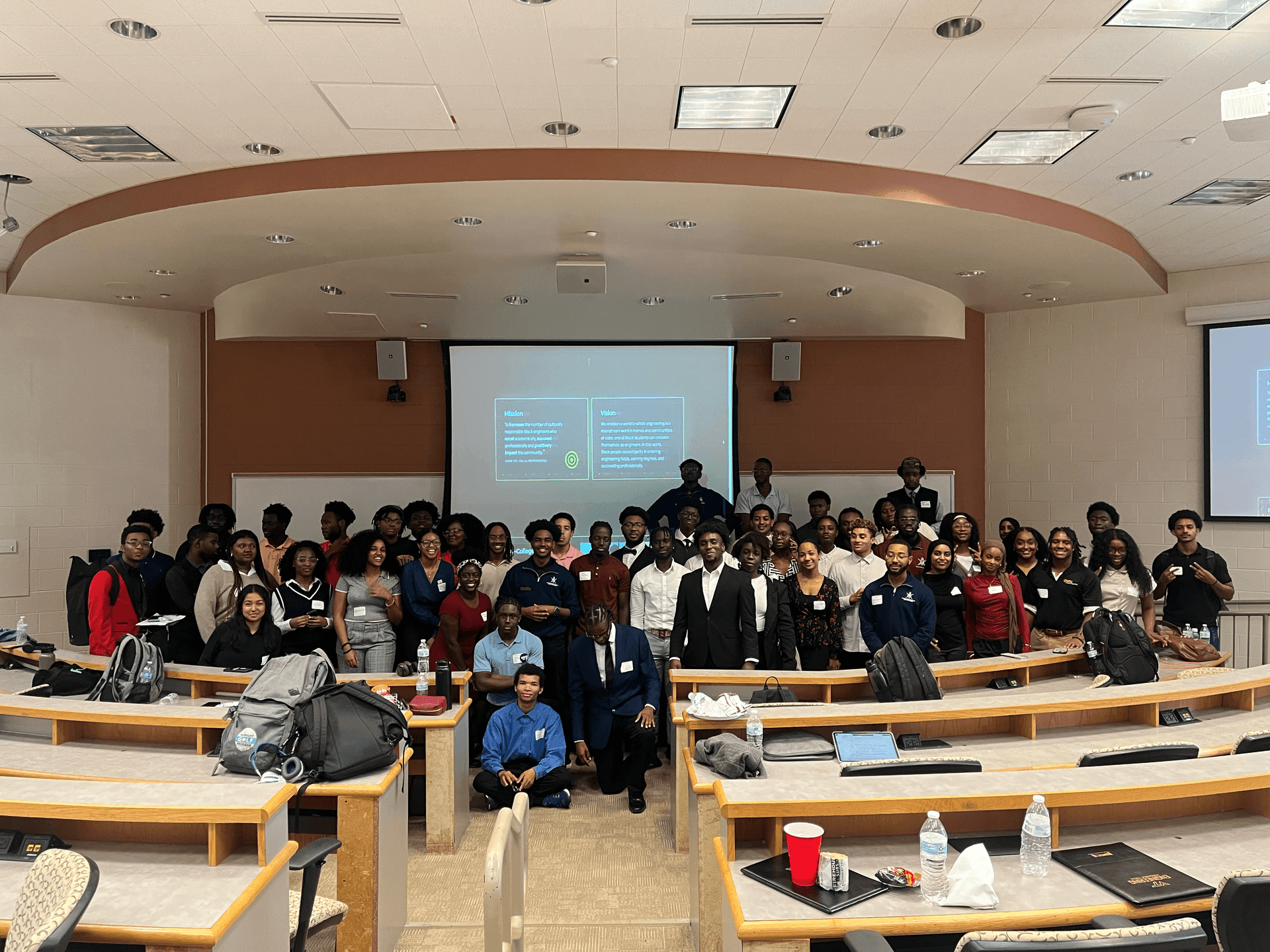 Large group of students posing in a lecture hall with a presentation screen and desks in the background.